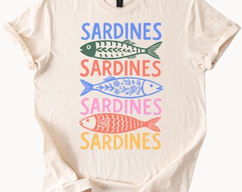May include: Cream-colored t-shirt featuring the word "SARDINES" stacked three times with colorful fish illustrations. The fish are green, blue, and red, with decorative patterns. The text and fish designs are in blue, pink, orange, and green.