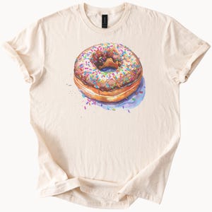 Puede incluir: Camiseta color crema con una ilustración de acuarela de una rosquilla. La rosquilla tiene glaseado de chocolate y está cubierta de chispas de colores. La camiseta es de material suave y tiene cuello redondo clásico.