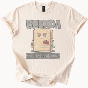 May include: A cream-colored t-shirt featuring a graphic of a beige towel character with a tired expression, wearing a red name tag that says BRENDA. Above the character is the text BRENDA, and below is the text SHE HAS SEEN THINGS.