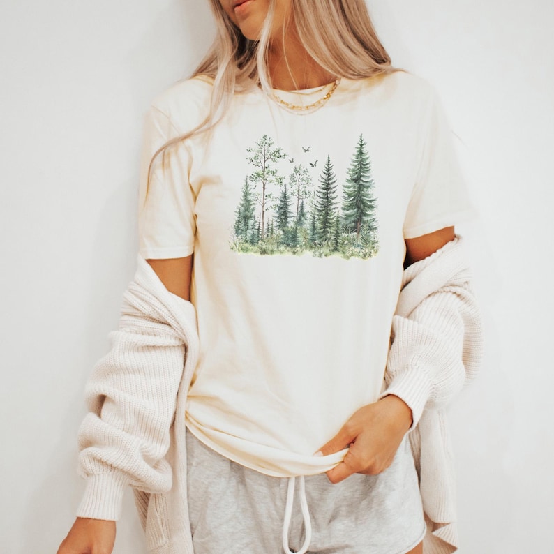 May include: Cream-colored t-shirt featuring a watercolor illustration of a forest scene with various trees and birds. The shirt is paired with gray shorts and a cream-colored cardigan. The overall aesthetic is casual and nature-inspired.