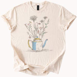 Può includere: T-shirt color crema con un'illustrazione stravagante di una teiera piena di fiori di campo. La teiera è bianca con dettagli blu e gialli. I fiori di campo sono raffigurati in tonalità di rosa, verde e grigio. La maglietta è in materiale morbido.