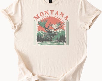 May include: Cream-colored t-shirt featuring a graphic of a moose in a lake with mountains and trees. The word "MONTANA" is arched above the moose in red letters. The design has a vintage, outdoorsy aesthetic.
