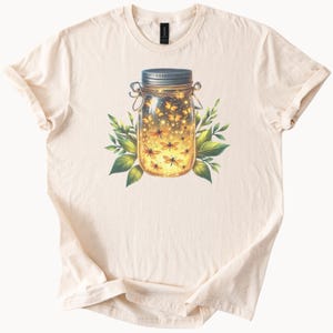 Op de afbeelding: Crèmekleurig T-shirt met een levendige illustratie van een weckpot vol gloeiende vuurvliegjes. De pot heeft een zilveren deksel en is omgeven door groene bladeren. De vuurvliegjes zijn afgebeeld in tinten geel en oranje.