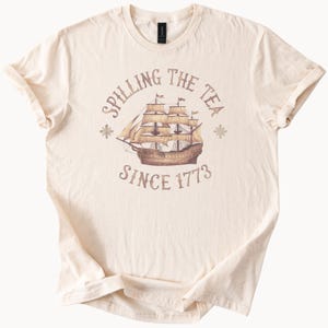May include: A beige t-shirt with a graphic of a ship and the text "Spilling the Tea Since 1773".