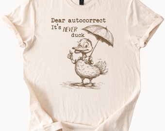 May include: A beige t-shirt with a brown illustration of a duck holding an umbrella. The text "Dear autocorrect It's NEVER duck" is printed on the shirt.