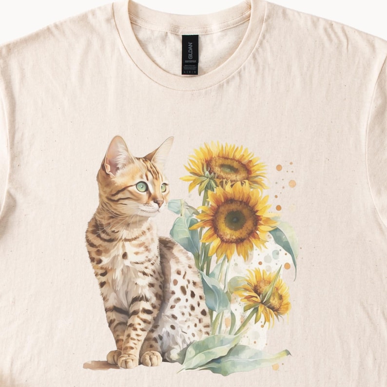 May include: A beige t-shirt featuring a watercolour illustration of a brown and white spotted cat sitting in front of a bunch of yellow sunflowers.