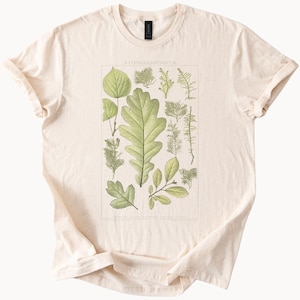 May include: Cream-colored t-shirt featuring a botanical illustration of various leaves and plants. The graphic is framed and includes the text "NATURKUNDLICHES." The shirt has short sleeves and a crew neck.