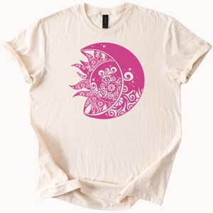 May include: Cream-colored t-shirt featuring a vibrant pink design of a crescent moon and sun. The sun and moon are intricately detailed with swirling patterns. The shirt has short sleeves and a crew neck.