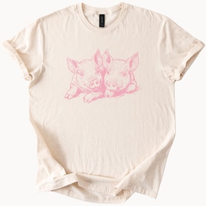 Könnte beinhalten: Ein cremefarbenes T-Shirt mit einer rosa Illustration von zwei Ferkeln, die sich gegenüberstehen.