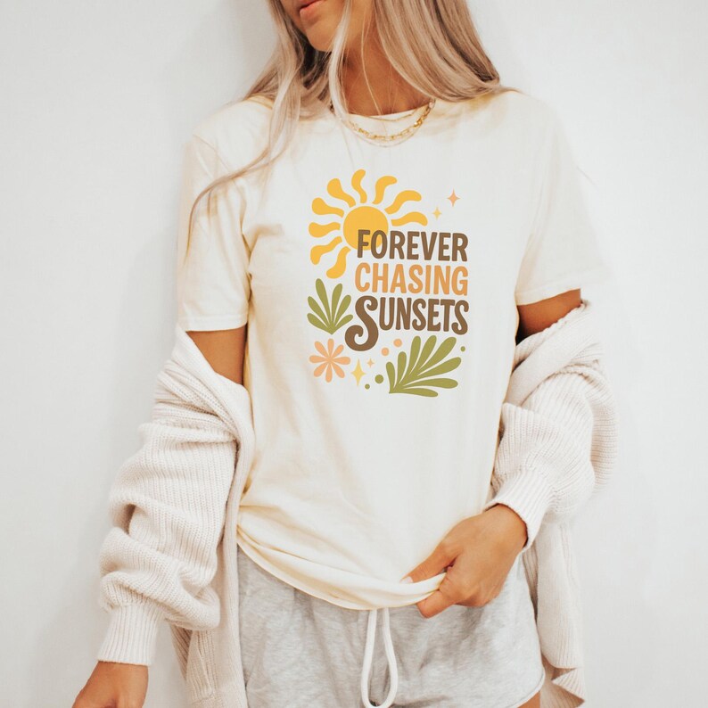 May include: Cream-colored t-shirt with the text "FOREVER CHASING SUNSETS" in brown, orange, and green. The design includes a sun, flowers, and leaves. The shirt is short-sleeved.