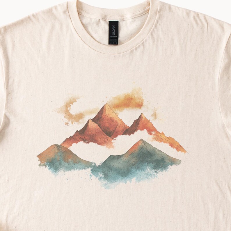 Op de afbeelding: Een beige t-shirt met een aquarel-illustratie van bergen en wolken. De bergen zijn bruin en oranje, en de wolken zijn blauw en groen.