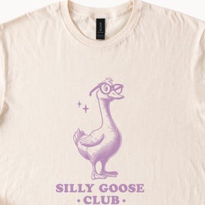 Puede incluir: Una camiseta beige claro con una ilustraci&oacute;n morada de un ganso con gafas. El ganso mira hacia la derecha. El texto "Silly Goose Club." est&aacute; impreso debajo del ganso.