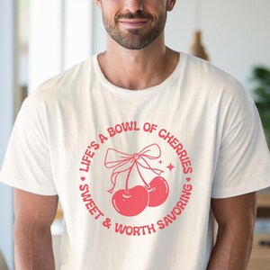 May include: White t-shirt with a pink graphic design of two cherries with a bow on top. The text reads "Life's a bowl of cherries - sweet & worth savoring."