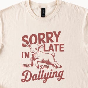 May include: Cream-colored t-shirt with a graphic of a lamb jumping and the text "SORRY I'M LATE I WAS Dilly Dallying" in maroon. The shirt has a crew neck and short sleeves.