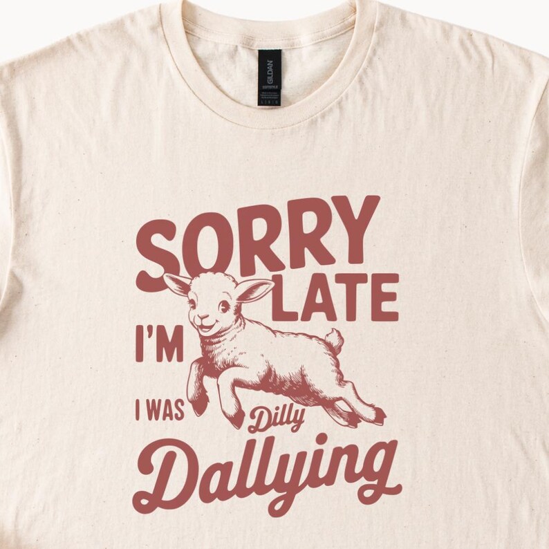 May include: Cream-colored t-shirt with a graphic of a lamb jumping and the text "SORRY I'M LATE I WAS Dilly Dallying" in maroon. The shirt has a crew neck and short sleeves.