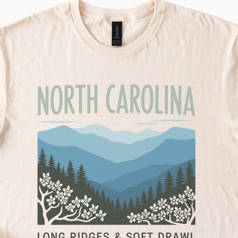 May include: Cream-colored t-shirt featuring the text "NORTH CAROLINA" in light green above a graphic of blue mountain ranges and trees. The design includes white flowering branches at the bottom. The shirt is a Gildan brand.