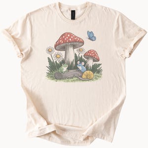 Könnte beinhalten: Cremefarbenes T-Shirt mit einer skurrilen Illustration einer Waldszene. Das Design zeigt rot gekappte Pilze mit weißen Punkten, Gänseblümchen, einen Frosch, eine Maus, eine Schnecke und einen blauen Schmetterling. Der Aufdruck befindet sich auf einem weichen, lässigen T-Shirt.