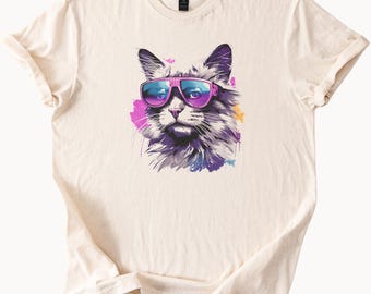 May include: A cream colored t-shirt with a graphic of a cat wearing sunglasses. The cat is surrounded by colorful paint splatters.