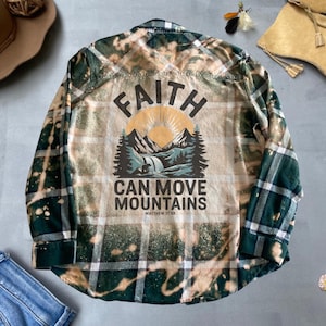 Op de afbeelding: Een groen en wit geruit flanellen overhemd met een gebleekt ontwerp. De achterkant van het overhemd heeft een afbeelding van een berglandschap met een zon en de tekst "FAITH CAN MOVE MOUNTAINS MATTHEW 17:20".