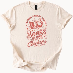 May include: Cream-colored t-shirt with a red graphic. The graphic includes the text "A woman cannot survive on books alone, she also needs chickens" with a stack of books and a rooster illustration.