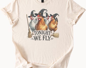 May include: Cream-colored t-shirt featuring a whimsical Halloween design. The graphic depicts four chickens wearing witch hats and holding brooms, with the text "TONIGHT WE FLY" below. A fun and festive apparel item.