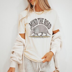 May include: A cream-colored short-sleeve t-shirt featuring a graphic of an armadillo walking on cracked ground. Above the armadillo, the text reads "MIND THE ROAD" in a dark gray arched font. The shirt is worn with a cream-colored cardigan.