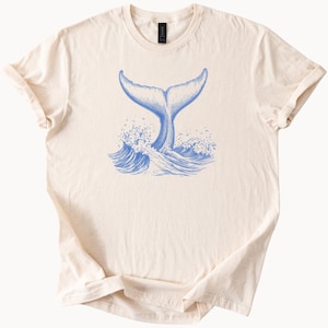 以下が含まれることがあります： クリーム色のTシャツに、青い海の波しぶきの中に、クジラの尾のイラストが描かれています。