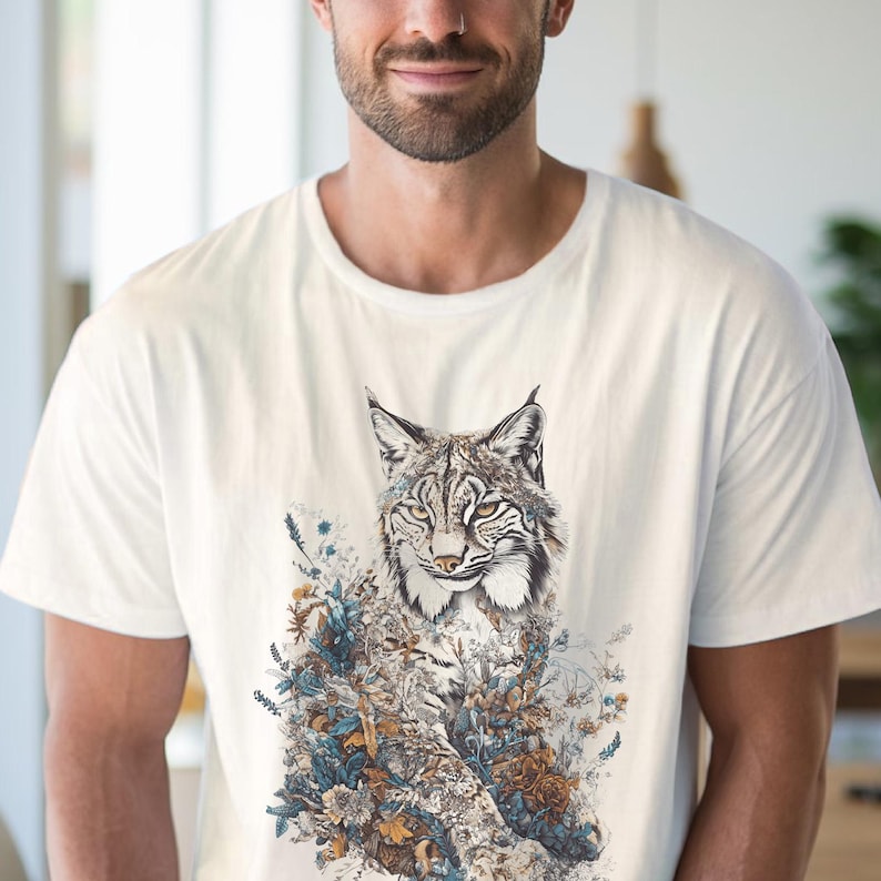 May include: White t-shirt featuring a detailed illustration of a lynx with floral accents. The lynx has a patterned coat and is surrounded by blue, brown, and white flowers. The shirt is a classic crew neck style.