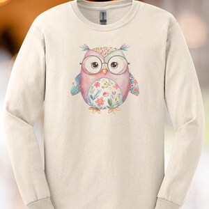 Peut inclure: T-shirt à manches longues de couleur crème avec un motif de hibou fantaisiste. Le hibou est rose, bleu et orné de motifs floraux, portant des lunettes. Le motif est centré sur le t-shirt, offrant une esthétique ludique et artistique.