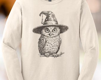 May include: Cream-colored long-sleeve shirt featuring a detailed illustration of an owl wearing a witch's hat. The owl is perched on a branch, with intricate feather and hat details. The shirt has a classic crew neck and long sleeves.
