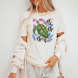 May include: Cream-coloured t-shirt featuring a vibrant sea turtle design with yellow and green details, surrounded by colourful fish and blue foliage. The graphic tee is a comfortable, casual wear option.