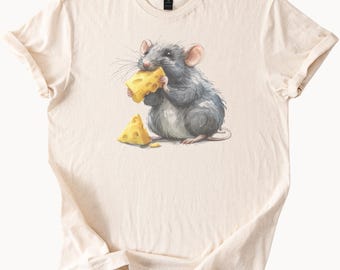 May include: Cream-colored t-shirt featuring a watercolor illustration of a gray rat eating a wedge of yellow cheese. A second cheese wedge sits on the shirt. The shirt has a crew neck and short sleeves.
