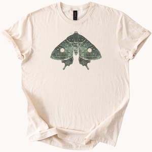 以下が含まれることがあります： クリーム色のTシャツ。モス（蛾）の精巧なイラストが描かれています。モスは緑色を基調とし、白いアクセントが施され、羽には2つの顕著な円形模様があります。Tシャツは、定番のクルーネックと半袖です。