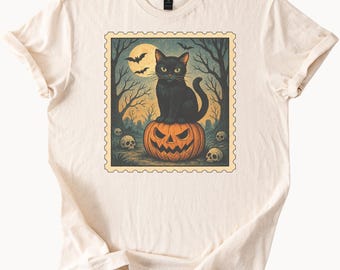 May include: A cream-coloured t-shirt with a Halloween graphic. The design shows a black cat on an orange jack-o'-lantern, with a full moon, bats, and trees in the background. Skulls are scattered around the base of the pumpkin.