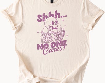 Camiseta gráfica divertida de esqueleto "Shhh, a nadie le importa" con cita sarcástica, estilo Halloween, para uso diario informal.