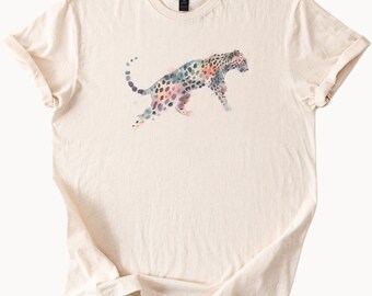 May include: Cream-colored t-shirt featuring a watercolor leopard design. The leopard is depicted in shades of blue, pink, and purple with black spots. The shirt has a classic crew neck and short sleeves, perfect for casual wear.