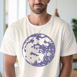 Op de afbeelding: Een wit T-shirt met een paarse maanprint. De maan is een gedetailleerde illustratie met kraters en een textuur.