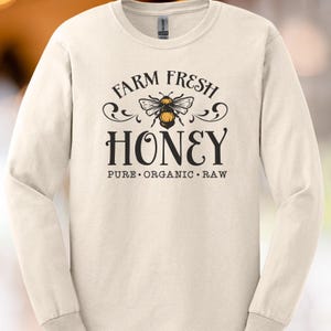 May include: Cream-colored long-sleeve shirt with a graphic that reads "FARM FRESH HONEY" in a vintage style. The design includes a bee illustration and the words "PURE ORGANIC RAW".