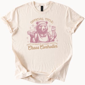 May include: Cream-colored t-shirt featuring a pink graphic of a bear holding a sandwich and a sock puppet. The text reads "OFFICIAL TITLE: SNACK DISTRIBUTOR/LAUNDRY OPERATOR/Chaos Controller."