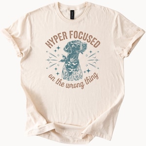 Op de afbeelding: Crèmekleurig T-shirt met een hondenillustratie met een bandana. De tekst luidt "HYPER FOCUSED on the wrong thing" in bruin en blauwgroen. Het ontwerp bevat sterren en lijnen rond de hond.