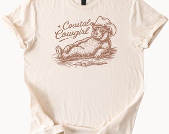 May include: Cream-colored t-shirt featuring a relaxed bear wearing a cowboy hat and sunglasses. The bear is lying down with the text "Coastal Cowgirl" above it. The design is in brown ink.