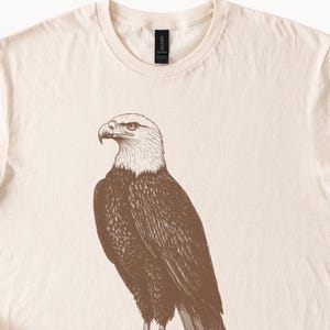 May include: A beige t-shirt with a brown illustration of a bald eagle on the front.