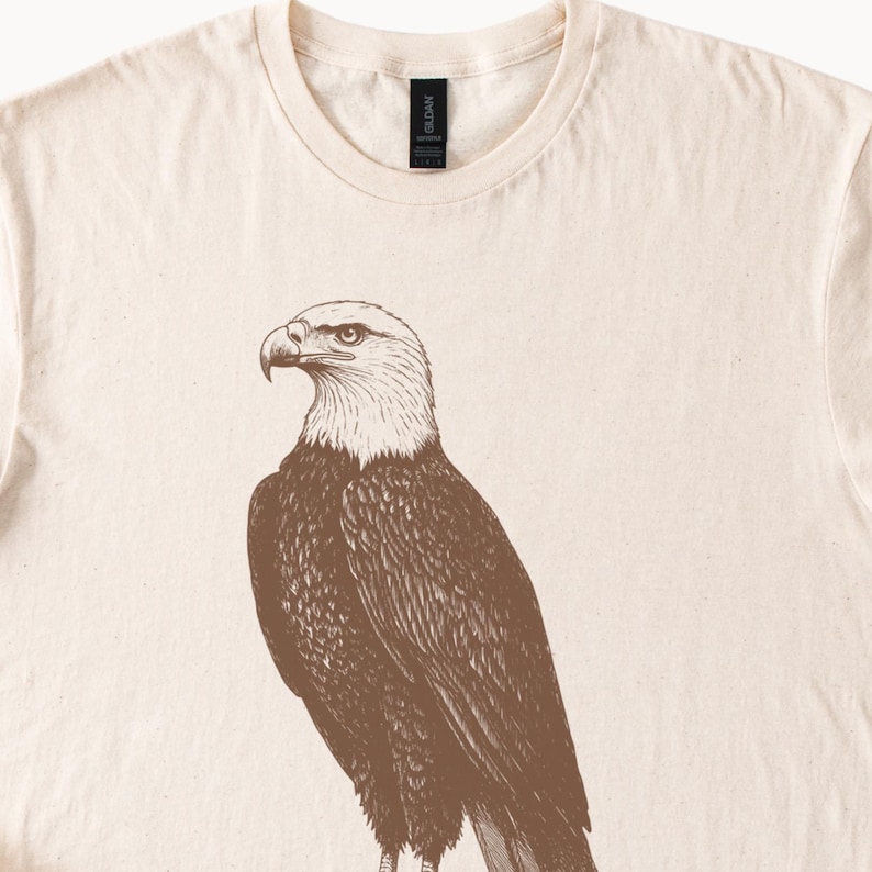 May include: A beige t-shirt with a brown illustration of a bald eagle on the front.