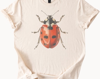 May include: Cream-colored t-shirt featuring a detailed illustration of a ladybug. The ladybug has a red shell with black spots and a black head. The shirt is made of soft material and has short sleeves.