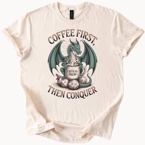以下が含まれることがあります： ベージュのTシャツに、緑色のドラゴンが「Critical Brew 1」と書かれたコーヒーカップを持っています。ドラゴンはサイコロの上に座っています。Tシャツには「Coffee First, Then Conquer」と書いてあります。