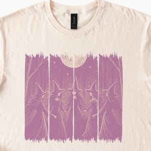 Pode incluir: Camiseta creme com uma ilustração roxa de cinco bruxas com chapéus pontudos e vestes, em pé em círculo sob uma lua cheia. O design tem um estilo vintage, de xilogravura, com linhas verticais que criam um efeito texturizado.