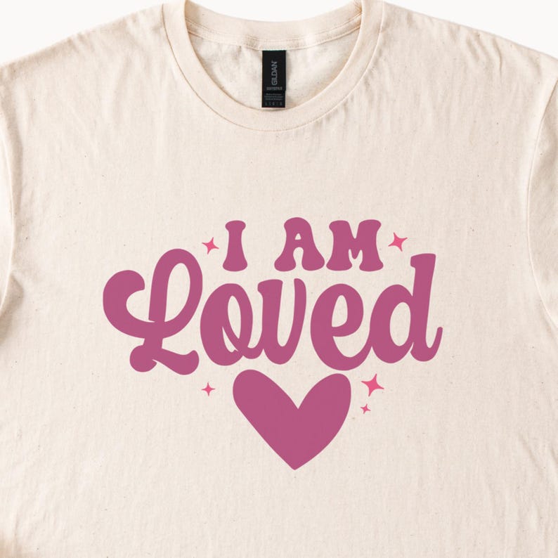 Puede incluir: Camiseta color crema con la frase "I AM Loved" en rosa intenso. La palabra "Loved" est&aacute; estilizada con un dise&ntilde;o de coraz&oacute;n. La camiseta tiene cuello redondo y mangas cortas.