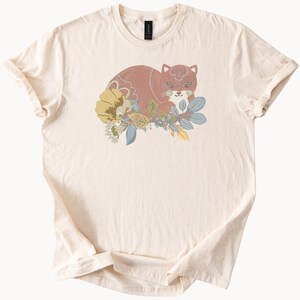 May include: A cream-coloured t-shirt with a graphic of a cat in brown tones, nestled amongst a floral design with yellow, blue, and green leaves. The t-shirt has a crew neck and short sleeves. The design is printed on a light-coloured fabric.