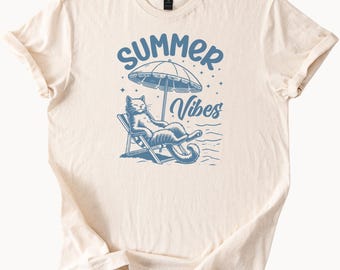 May include: Cream-colored t-shirt featuring a blue graphic of a cat relaxing in a beach chair under an umbrella. The words "Summer Vibes" are arched above the cat. The shirt has rolled-up sleeves.