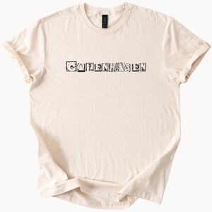 Puede incluir: Camiseta color crema con la palabra "COPENHAGEN" impresa en negro, con letras de bloque. La camiseta tiene cuello redondo y mangas cortas, con las mangas enrolladas. La tela parece suave y ligeramente texturizada.